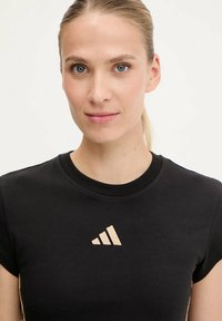 Maglietta atletica nera a maniche corte in materiale morbido con il logo Adidas beige sul petto. Collo rotondo con maniche risvoltabili.