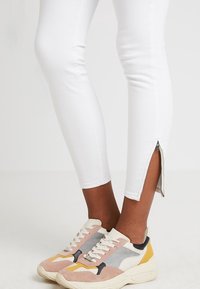 Jeans blancos hasta el tobillo con un detalle de cremallera lateral, combinados con zapatillas multicolores que tienen acentos en rosa, gris y amarillo.