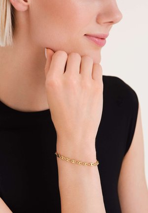 Bracelet en chaîne dorée avec des maillons ovales interconnectés, texture lisse, porté sur un poignet contre un haut sombre, design minimaliste.
