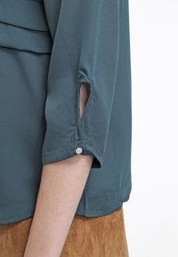 Blouse bleu sarcelle avec une coupe décontractée, des manches 3/4 avec des accents de bouton, et une texture lisse. La manche présente un petit détail de découpe.