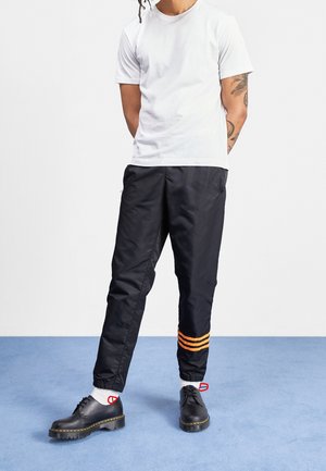 Schwarze Nyluhosen mit orangefarbenen Streifen an den Bündchen, kombiniert mit einem schlichten weißen T-Shirt und schwarzen Schuhen mit dicker Sohle.