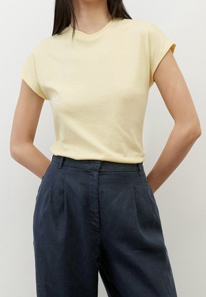 Vrouw draagt een lichtgeel T-shirt met korte mouwen, ingestopt in hoog getailleerde donkerblauwe geplooide broek met handen achter haar rug.