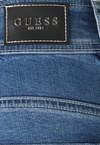 Blue denim fabric with a textured surface, featuring a black leather patch labeled "GUESS EST. 1981" with silver lettering and white stitching.