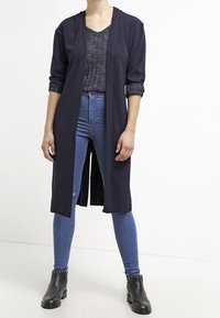 Lange marineblauwe cardigan met open voorkant en opgerolde mouwen, gecombineerd met een gestructureerd grijs topje en blauwe skinny jeans. Zwarte enkellaarsjes met studs.