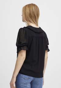 Sort bluse med prikker, pufærmer og flæset halsudskæring. Båret med blå denim jeans, set fra ryggen.