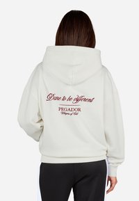 Sudadera crema con cordón y bolsillo canguro. La parte trasera presenta texto bordado en rojo: "Atrévete a ser diferente" y "PEGADOR, Susurros de Oro."