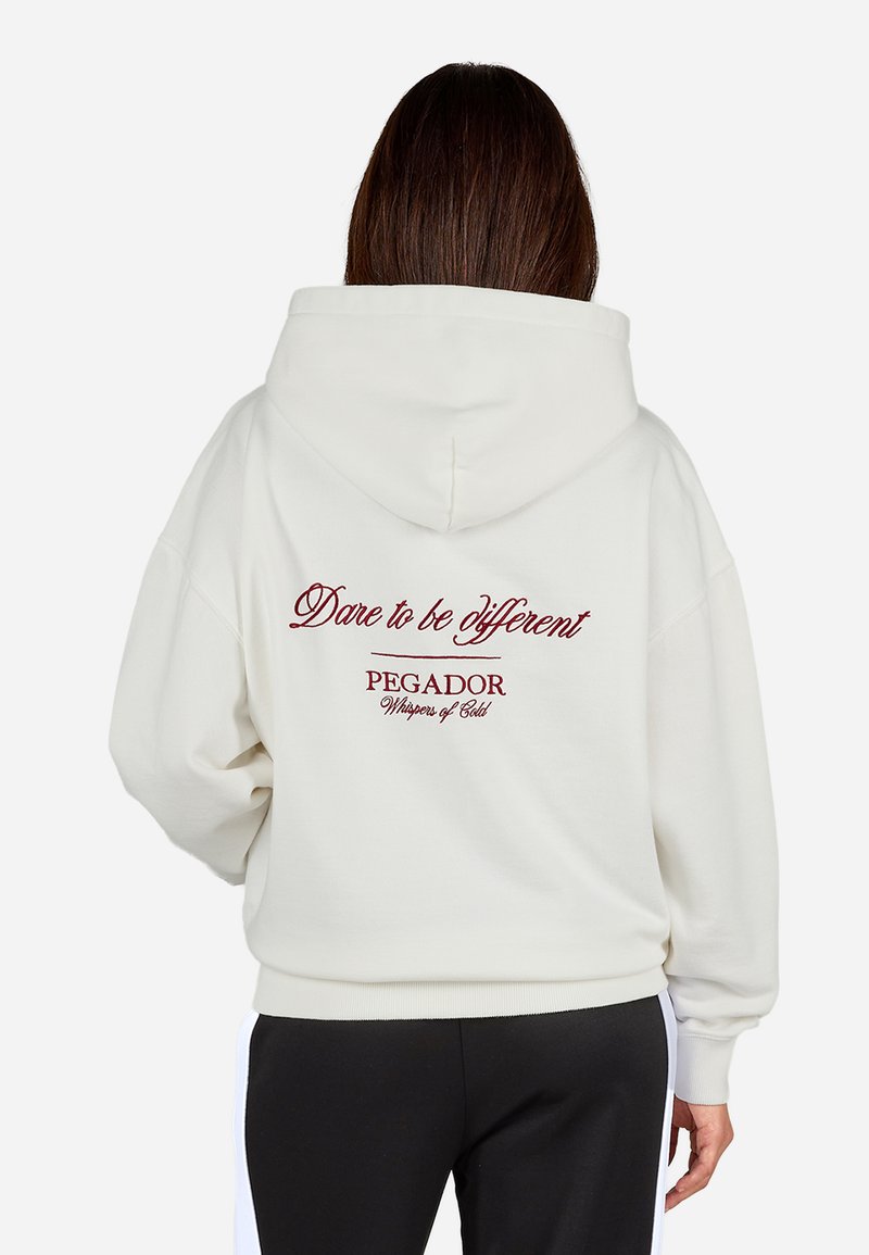 Sudadera crema con cordón y bolsillo canguro. La parte trasera presenta texto bordado en rojo: "Atrévete a ser diferente" y "PEGADOR, Susurros de Oro."