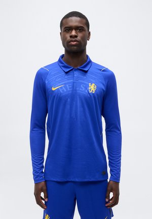 Mężczyzna ubrany w niebieską, długorękawową koszulkę piłkarską Chelsea FC oraz pasujące spodenki, stojący przodem na jednolitym tle.
