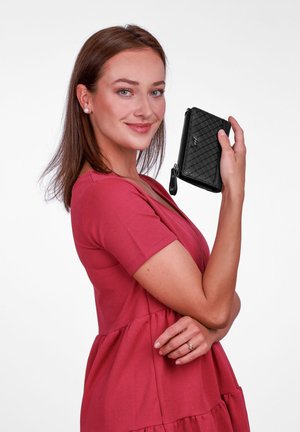 Black quilted wallet with zipper closure, featuring a textured pattern and a branded tag, held in hand against a plain white background.