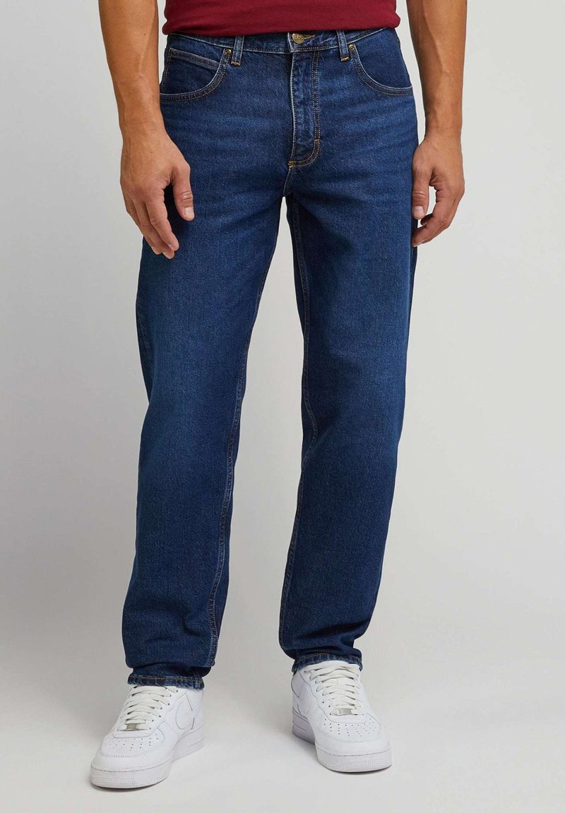 Mørkeblå denimjeans med regular fit, fem lommer og synlige sømdetaljer. Kombinert med hvite joggesko.