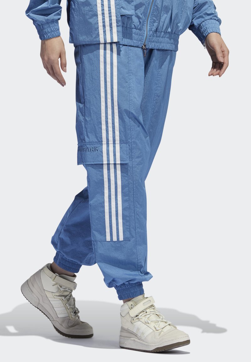 Lichtblauwe sportieve trainingsbroek met witte strepen, voorzien van elastische boorden en een cargozak. Gepaard met witte high-top sneakers.