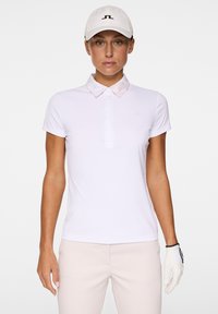 Biała koszulka polo z krótkim rękawem i subtelnym kołnierzykiem, z minimalistycznym logo. Połączona z beżowymi spodniami i białą czapką.