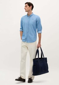 Lichtblauw overhemd met knopen, crèmekleurige broek, donkergekleurde schoenen, houdend een marineblauwe gestructureerde tote bag met meerdere zakken en dubbele handvatten.