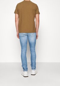 Brun kortærmet t-shirt parret med lyseblå skinny jeans og hvide sneakers, set fra ryggen mod en simpel hvid baggrund.