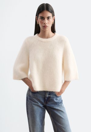 Femme aux longs cheveux foncés portant un pull crème duveteux à manches aux coudes et un jean bleu, debout les mains dans les poches face à un fond neutre.