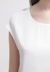 Blouse blanche avec un col arrondi, des manches courtes retroussées et des détails en dentelle transparente le long des coutures des épaules. Texture de tissu lisse.