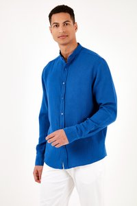 Man draagt een blauw overhemd met opstaande kraag en lange mouwen en witte broek, staand tegen een eenvoudige witte achtergrond.