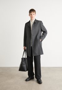 Grau melierter Wollmantel mit Reverskragen, zwei Knöpfen und seitlichen Taschen. Kombiniert mit einem hellen Hemd, schwarzen Hosen, schwarzen Schuhen und einer Tote-Tasche.