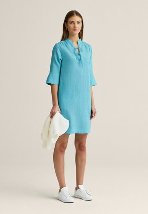 Kvinde iført en lys blå knælængde kjole med 3/4 ærmer, der holder en hvid sweater, stående mod en ensfarvet beige baggrund.