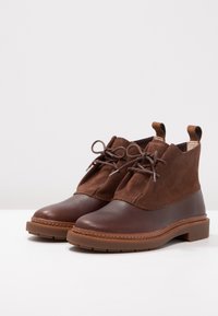 Botas tobilleras marrones hechas de cuero y ante, con punta redonda, diseño de cordones y tacón bajo tipo bloque con suela de goma texturizada.
