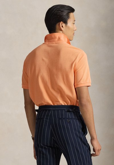Πόλο με κοντά μανίκια σε ανοιχτό πορτοκαλί με υφή, συνδυασμένο με παντελόνια κομμένα σε στυλ με λεπτές ρίγες σε ναυτικό μπλε, με πλαϊνές τσέπες και εφαρμοστή γραμμή.