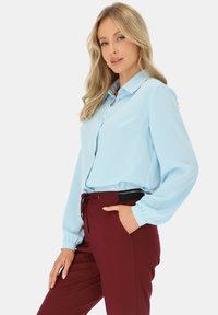 Kobieta z długimi blond włosami, ubrana w jasnoniebieską bluzkę z długim rękawem i bordowe spodnie, pozująca z jedną ręką w kieszeni na białym tle.