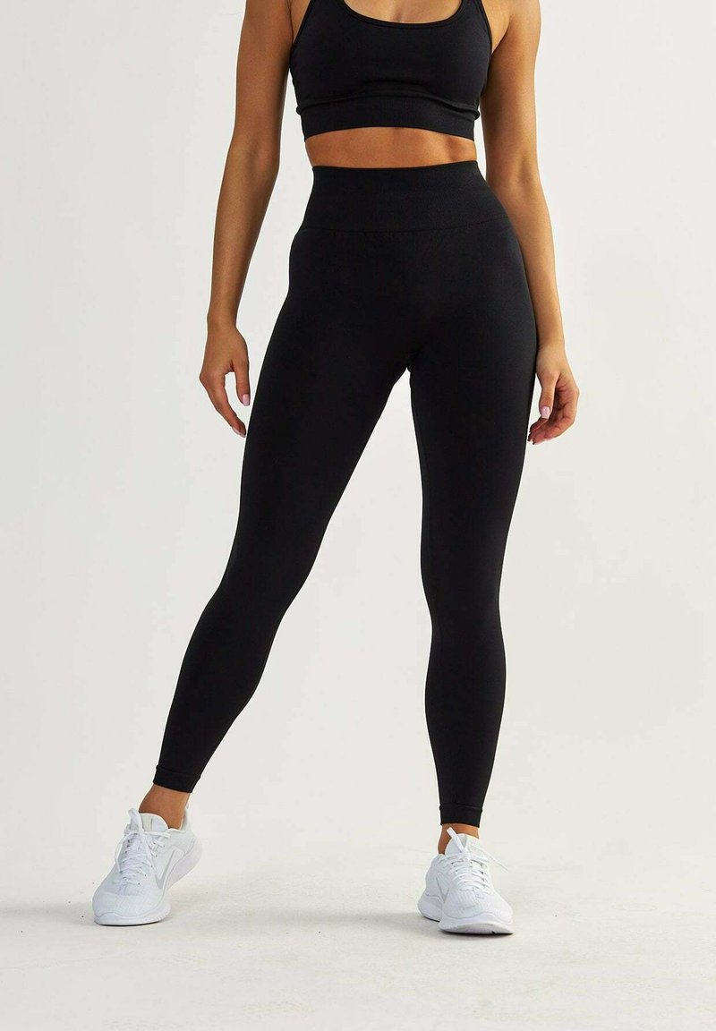 Leggings noirs taille haute en matériau extensible, avec une texture lisse et un design ajusté. Portés avec des chaussures de sport blanches.