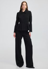 Žena v černém sako s peplum a širokými černými kalhotami, stojící na světlém šedém pozadí.