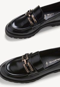 Mocassins en cuir noir avec semelles épaisses et détail décoratif de chaîne métallique sur le dessus, présentés sur un fond blanc.
