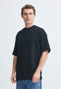 T-shirt noir surdimensionné à manches courtes, col rond, avec un détail de logo discret sur la poitrine. Porté avec un jean en denim bleu.