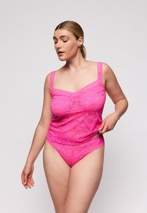 Vrouw die een felroze kanten lingerieset draagt, haar topje verstelt, staand tegen een effen lichte achtergrond.