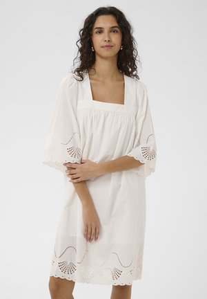 Femme aux cheveux bouclés et foncés portant une robe blanche à manches larges avec un bord en dentelle ajourée festonnée sur les manches et l'ourlet.