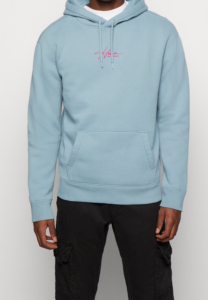 Ljusblå hoodie med ficka på framsidan, snörning och en rosa broderad logotyp på bröstet. Tillverkad av ett mjukt, strukturerat tyg.