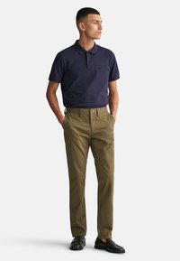 Camiseta polo azul marino con logo en relieve, combinada con pantalones chinos verde oliva y mocasines oscuros. El modelo está de pie con las manos en los bolsillos, sobre un fondo neutro.