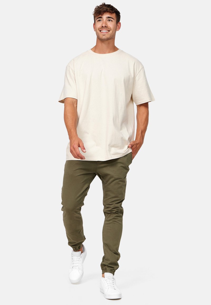 Giovane uomo che cammina, indossa una maglietta crema oversize, pantaloni da jogging verde oliva e sneakers bianche su uno sfondo grigio chiaro.