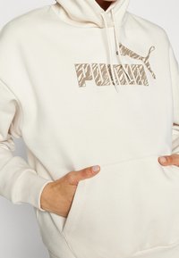 Bluza Puma w jasnym beżu, z kieszenią kangurzą, kapturem na sznurki i brązowym logo graficznym z akcentami w wzór zebry.