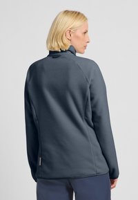 Giacca in fleece grigio scuro con colletto alto, maniche raglan e un'etichetta laterale. Tessuto morbido e testurizzato con una vestibilità relaxed e cuciture lisce.