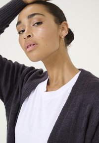 Cardigan lavorato a maglia di un grigio scuro con una texture a coste, indossato sopra una camicia bianca. Il cardigan presenta ampie revers e un tessuto morbido.