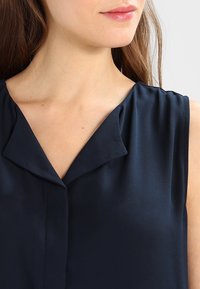 Granatowa bluzka bez rękawów o miękkiej, gładkiej fakturze i dekolcie w serek. Posiada subtelne plisy na ramionach i zapięcie na guziki z przodu.