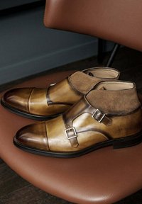Bottines en cuir marron avec un bout pointu, des accents en daim, deux boucles et une finition lisse, reposant sur une chaise en cuir marron.