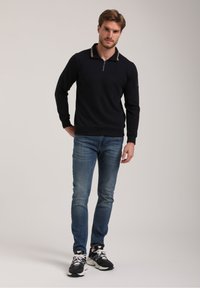 Marineblauer Hoodie mit Reißverschluss und strukturiertem oberen Teil, kontrastierendem Kragen, getragen mit hellblauen Jeans und schwarzen Sportschuhen.