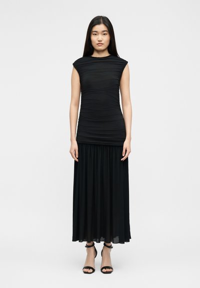 Γυναίκα που στέκεται όρθια, φοράει μαύρο αμάνικο φόρεμα με ρελιά και μακριά πιετέ φούστα, συνδυασμένο με μαύρες γόβες με λουράκι στον αστράγαλο.