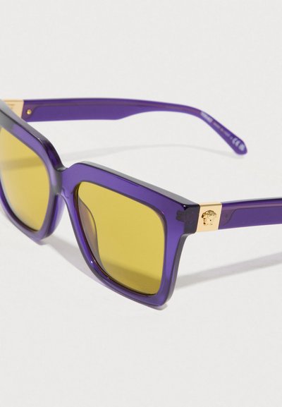 Μωβ διαφανή γυαλιά ηλίου με κίτρινους φακούς και ένα μικρό χρυσό έμβλημα στους βραχίονες, σε λευκό φόντο.