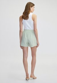 Shorts tissés vert clair avec une taille élastique, associés à un haut sans manches blanc. La mannequin se tient de dos face à la caméra.
