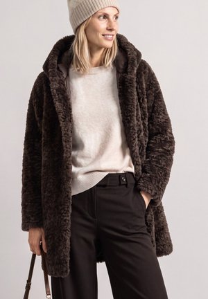 Femme portant un manteau en fausse fourrure marron, un pull crème, un bonnet beige en maille et un pantalon noir, tenant un sac à main marron de la main gauche.