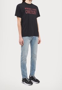 Černé bavlněné tričko s červeným nápisem "VERSACE JEANS COUTURE." V kombinaci se světle modrými džíny a černými teniskami s akcentovými detaily.