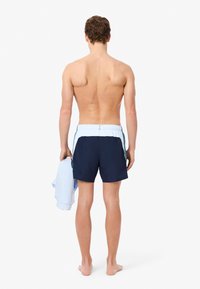 Marine zwempants met elastische tailleband, lichtblauwe accenten aan de zijkanten. Blootvoetse persoon die een lichtblauw handdoek vasthoudt, gezien van achter.