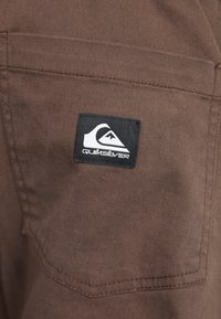 Barna szövetzseb fekete Quiksilver márkacímkével, rajta fehér hullám és hegyi logó.