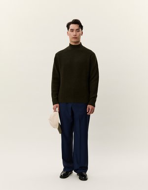 Jersey de punto verde oscuro con cuello alto, combinado con pantalones de marina y zapatos negros. El modelo sostiene una gorra beige en una mano.