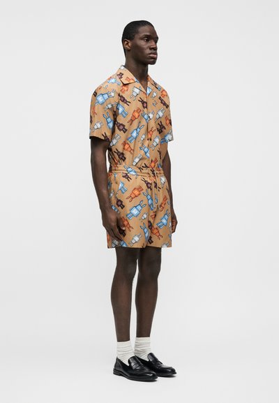  Ολόσωμο σορτς με κοντά μανίκια και κουμπιά, σε καφέ ύφασμα με πολύχρωμα παιδικά σχέδια μαχαιροπήρουνων. Ελαστική μέση με κορδόνι. Φορεμένο με μαύρα loafers.